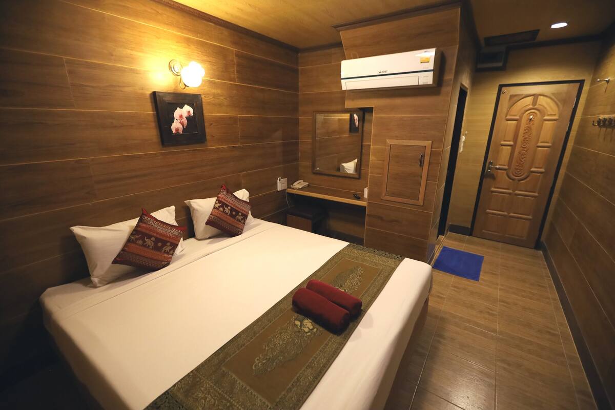 A hotel room with a wooden floor, a bed with white bedding, a red towel, a wall-mounted air conditioner, a mirror, and a door with a handle.
