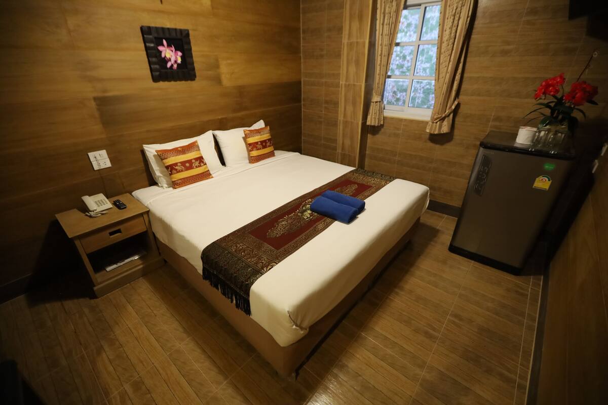 A hotel room with a wooden floor, a bed with a red and gold blanket, a nightstand with a phone, and a refrigerator with flowers.