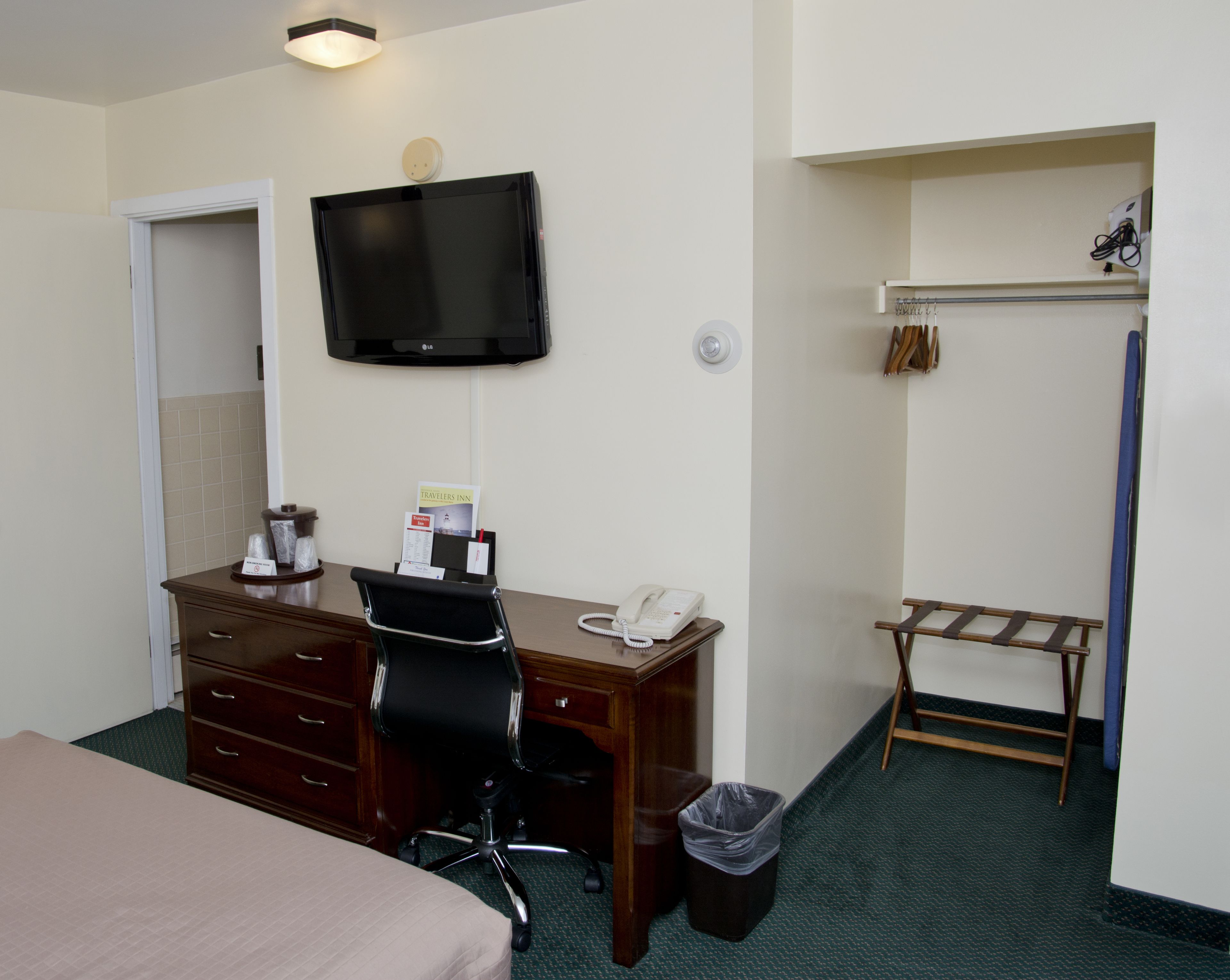 A hotel room with a bed, a desk with a telephone, a trash bin, a chair, a television mounted on the wall, and a coat rack with a coat.