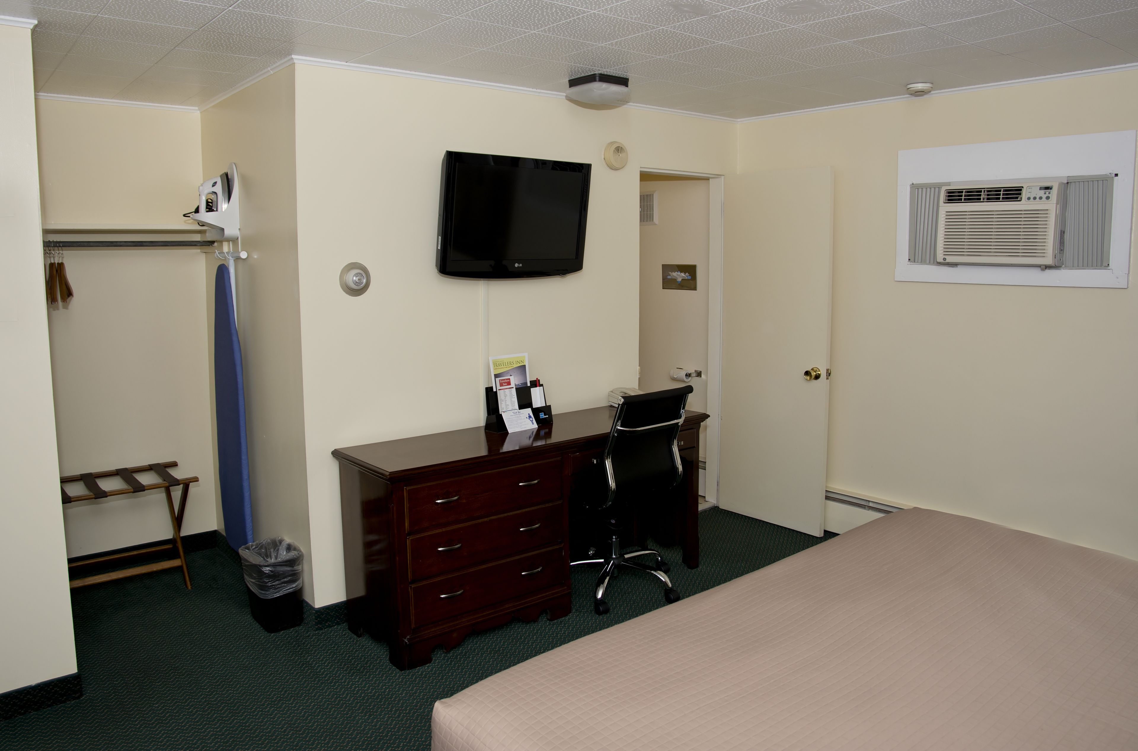 A hotel room with a bed, a desk with a chair, a television mounted on the wall, a coat rack, and an air conditioning unit.