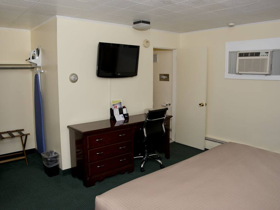A hotel room with a bed, a desk with a chair, a television mounted on the wall, a coat rack, and an air conditioning unit.