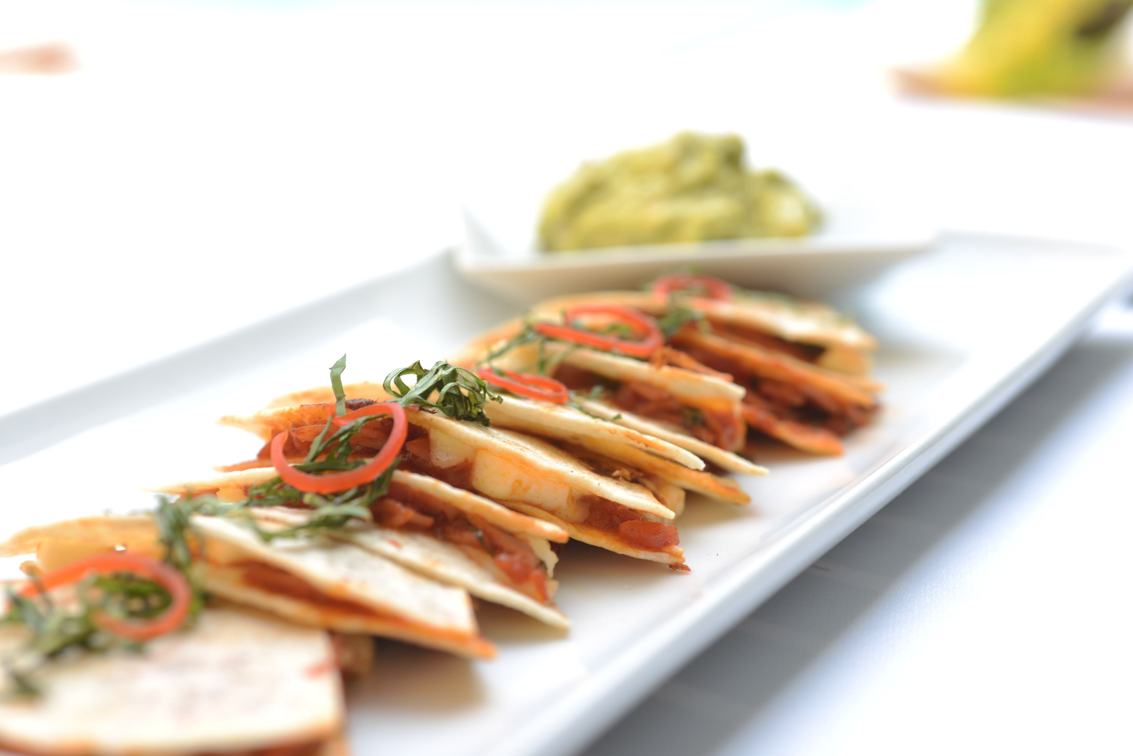 A white rectangular plate with a row of quesadillas garnished with red peppers and herbs, and a small bowl of guacamole in the background.