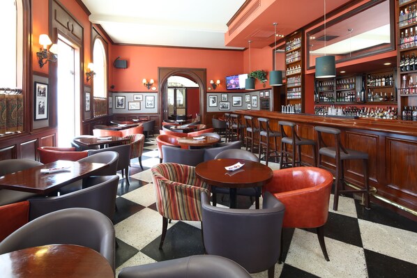 A bar area with multiple seating options including bar stools and round tables, a checkered floor pattern, and a variety of bar glasses and bottles on shelves. - LP Los Portales Hotel Piura (Piura)