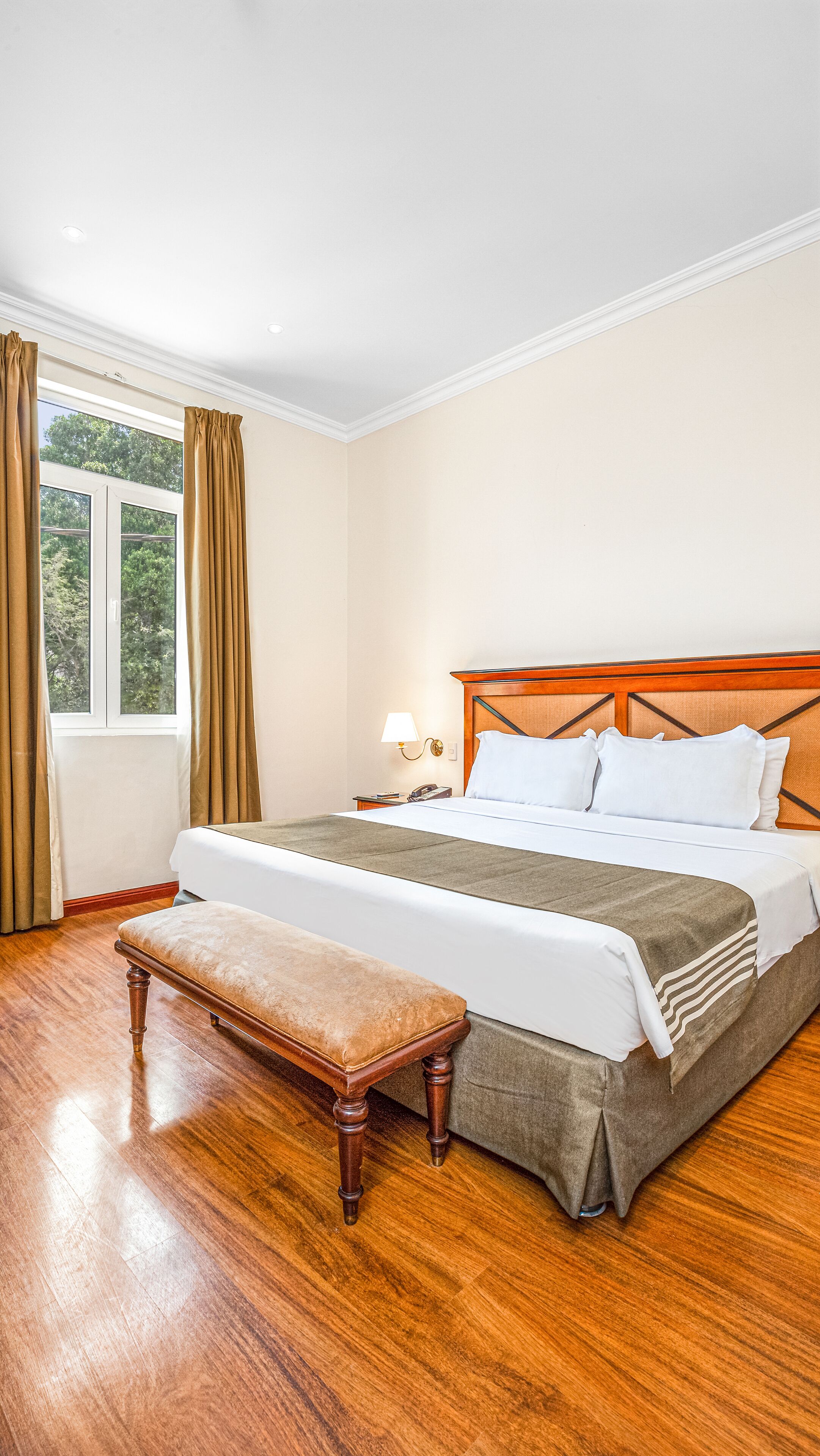 Habitación de hotel con una cama grande, un banco de madera, una mesita de noche con lámpara y una ventana con cortinas.