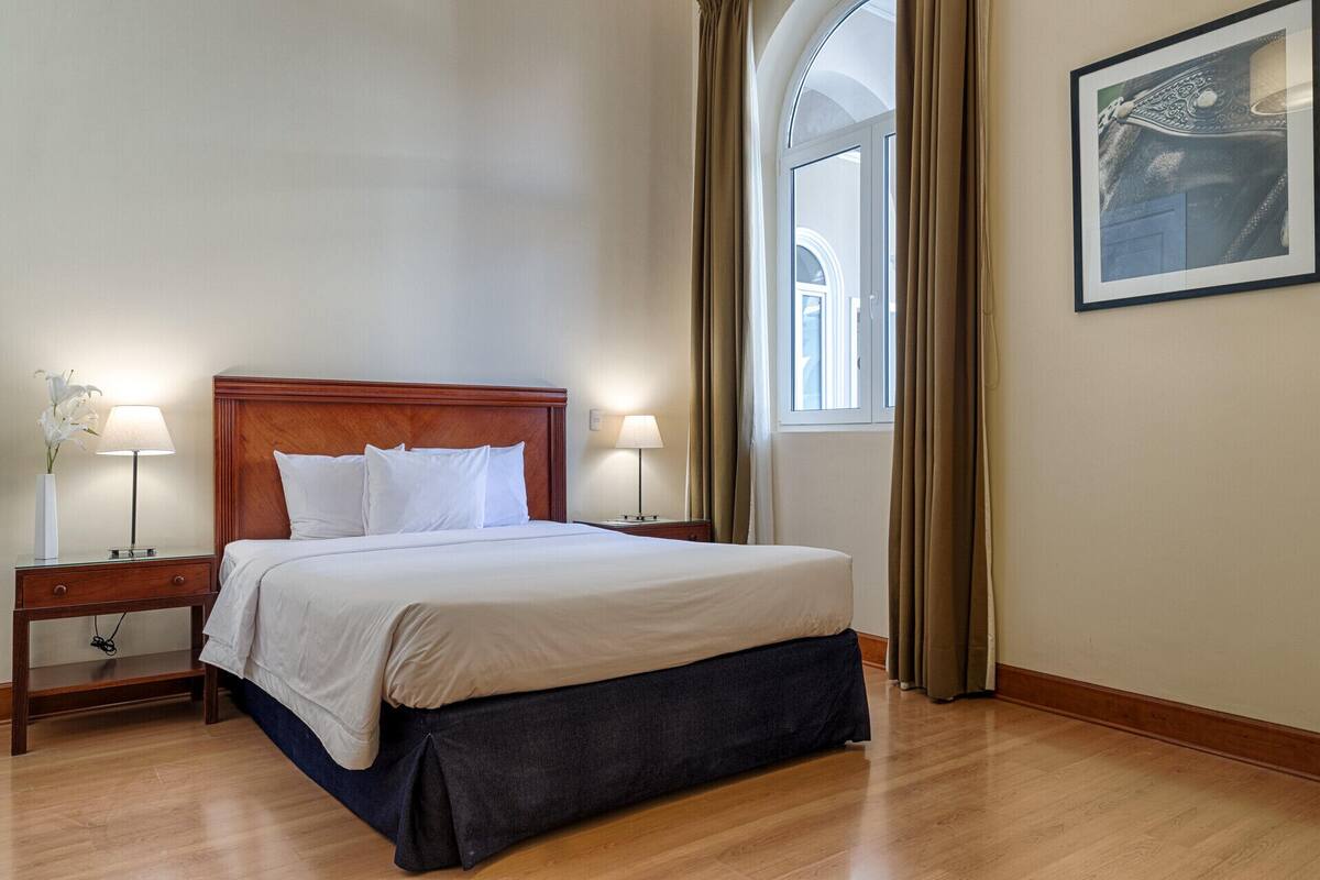 Une chambre d’hôtel avec une tête de lit en bois, deux tables de chevet avec des lampes, un grand lit avec une literie blanche, une fenêtre avec des rideaux et un tableau encadré au mur.