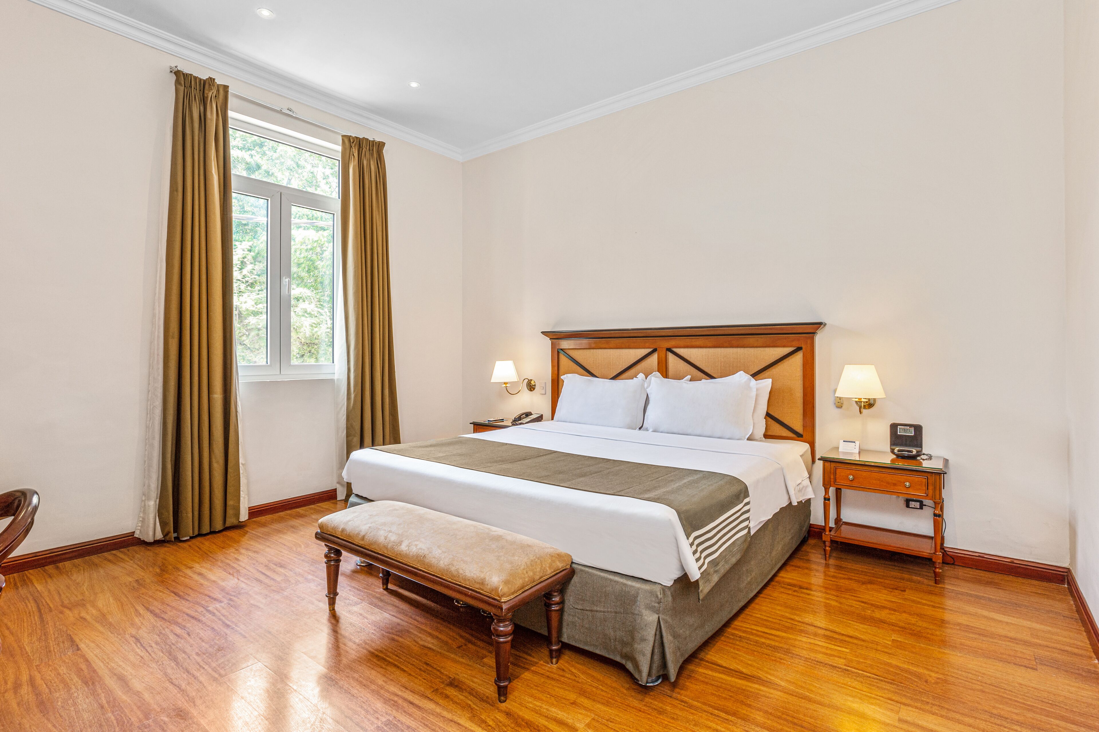 A hotel room with a large bed, wooden floor, a bench, two bedside tables with lamps, and a window with curtains.