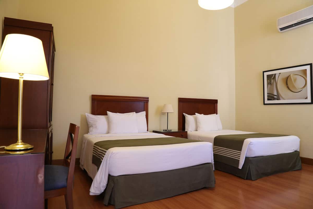 Une chambre d’hôtel avec deux lits, un bureau, une chaise, une lampe et un tableau encadré au mur.