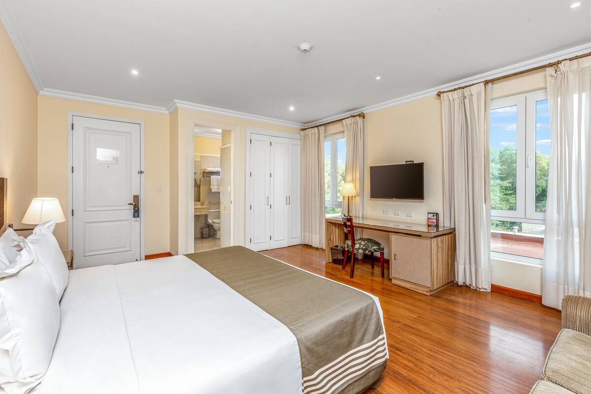 Une chambre d’hôtel avec un grand lit, un bureau avec une télévision et une vue sur la verdure environnante.