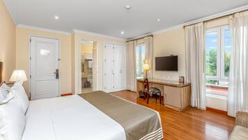 A hotel room with a large bed, a desk with a TV, and a view of greenery outside.