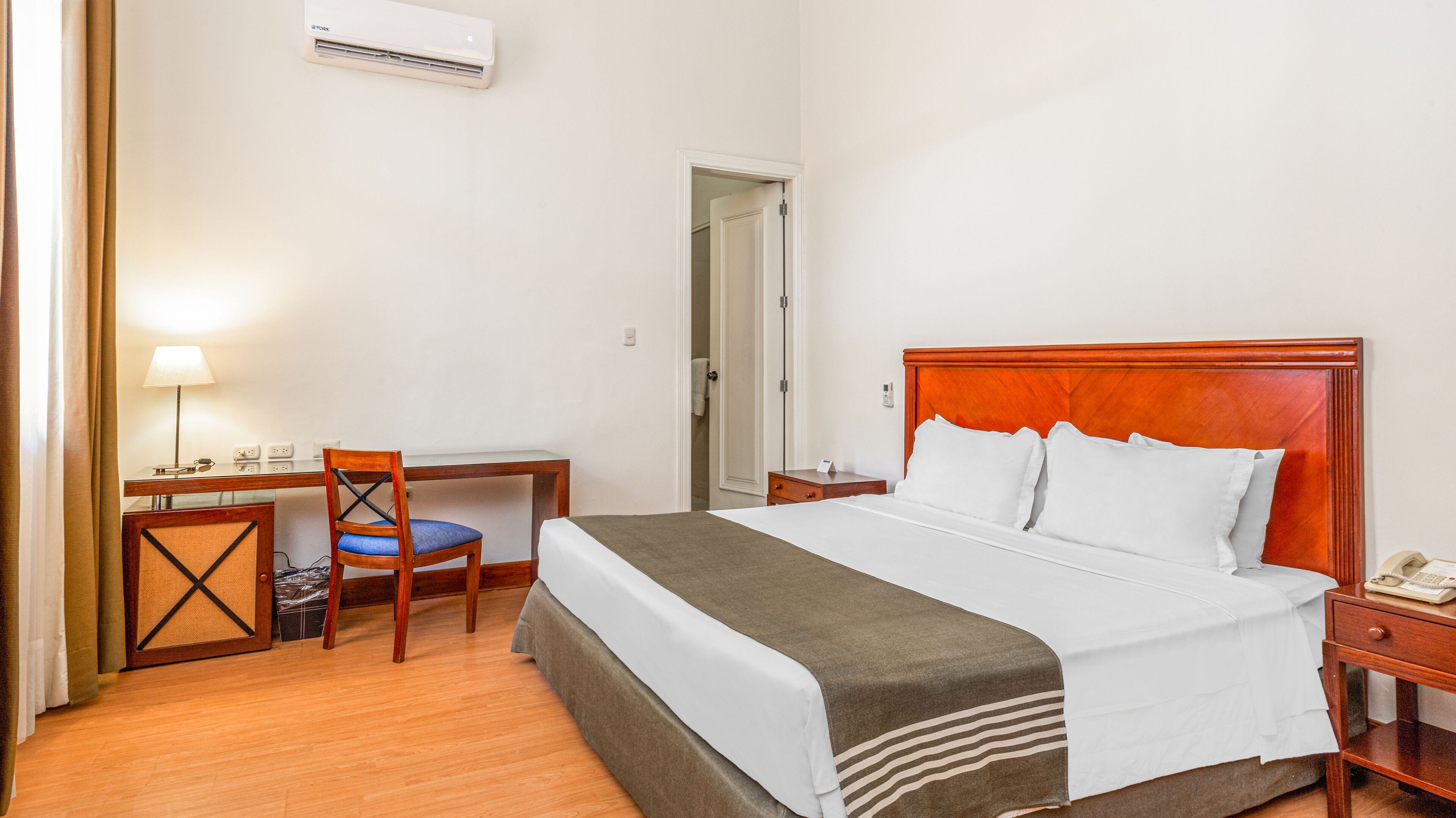 Une chambre d’hôtel équipée d’un lit, d’une table de chevet, d’une lampe, d’une chaise, d’un bureau et d’un appareil de climatisation.