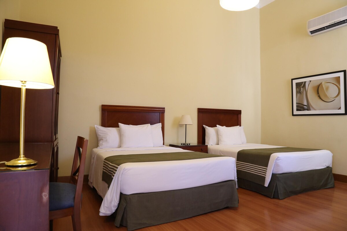 Habitación de hotel con dos camas, un escritorio con lámpara, una silla y un cuadro en la pared.