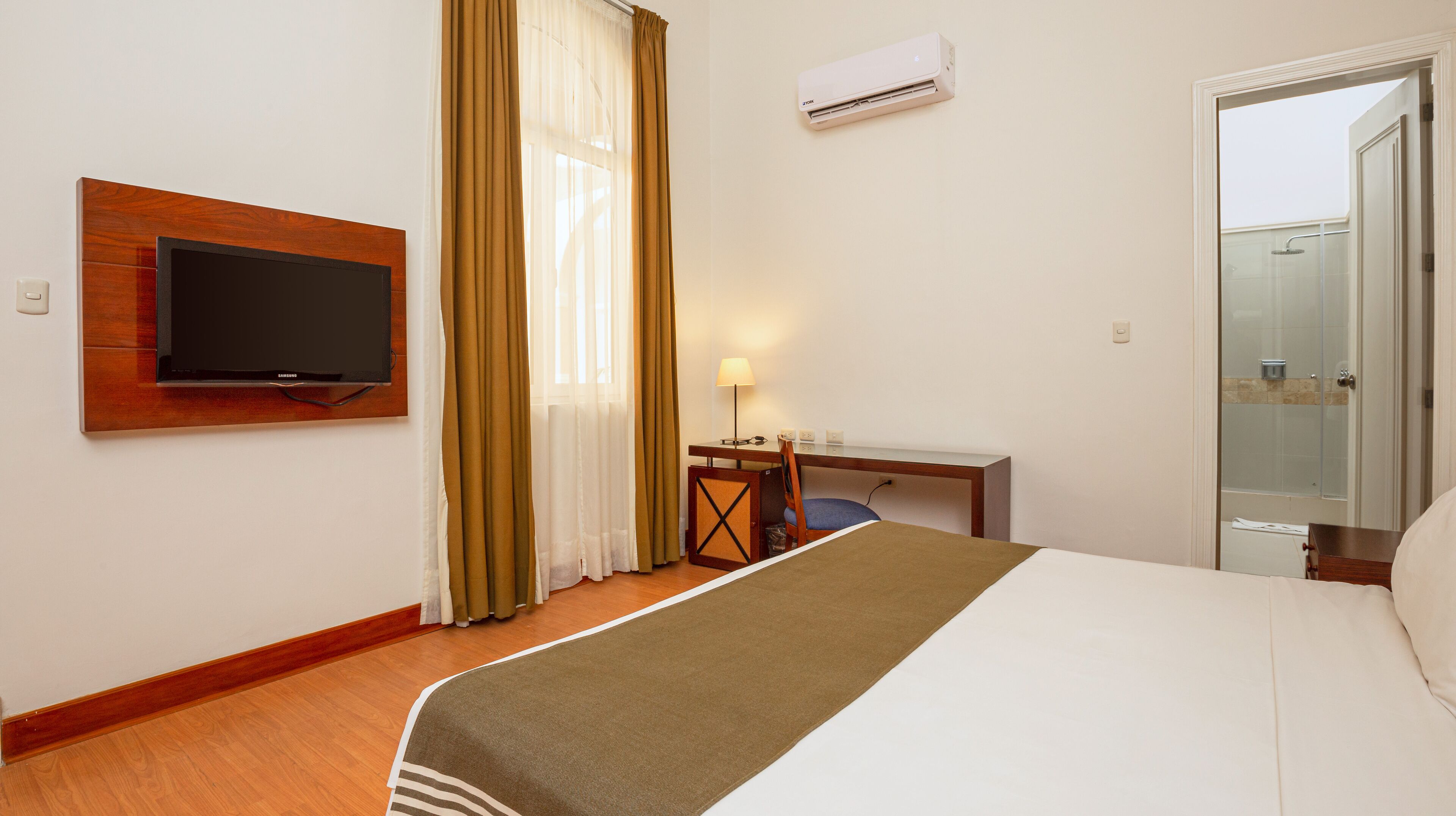 A hotel room with a bed, a desk, a chair, a television, a lamp, and an air conditioning unit.