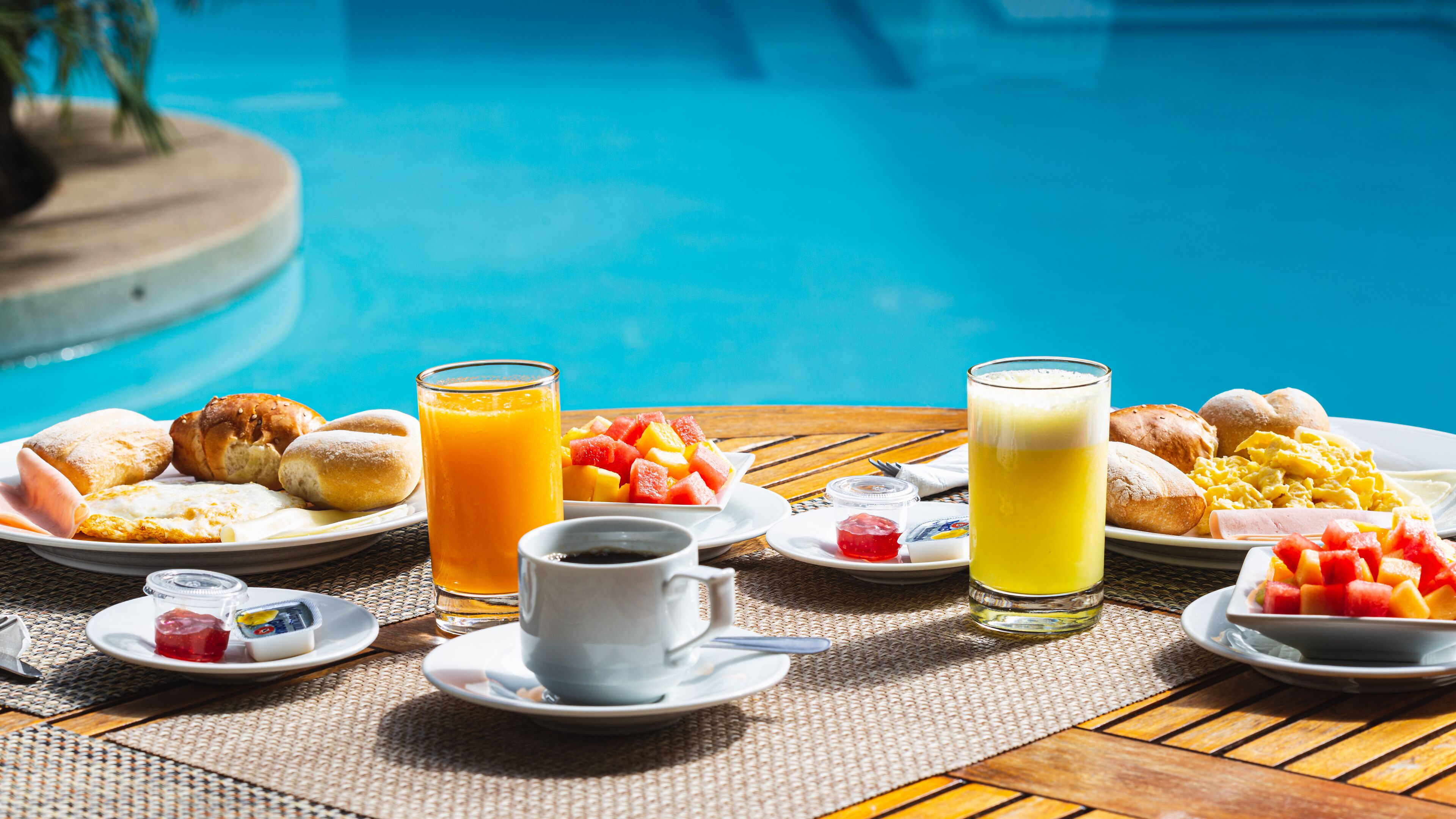 Desayuno junto a una piscina, con platos de comida, vasos de jugo y una taza de café.