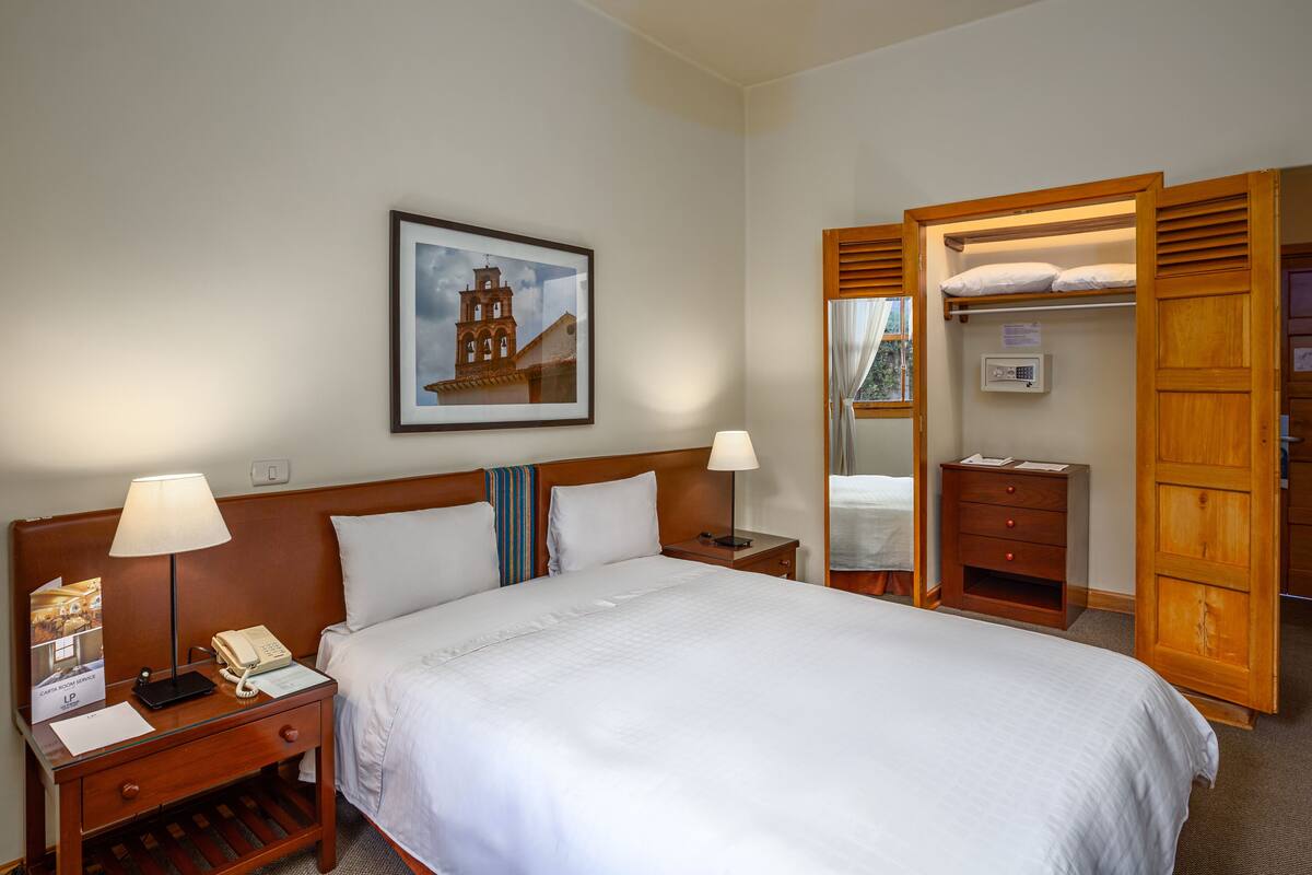 A hotel room with a large bed, two bedside tables with lamps, a wooden nightstand, a framed picture on the wall, and a wooden wardrobe.