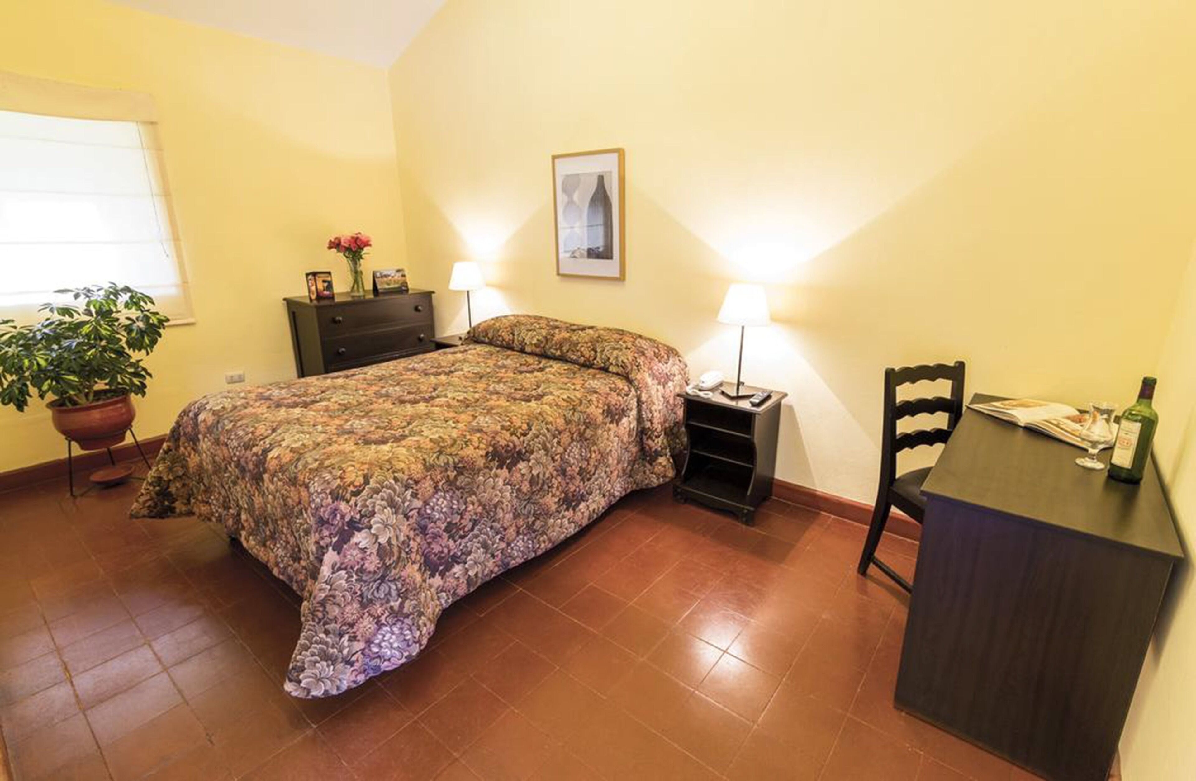 A bedroom with a floral bedspread, a bedside table with a lamp, a chair, a small table with a bottle, and a potted plant.