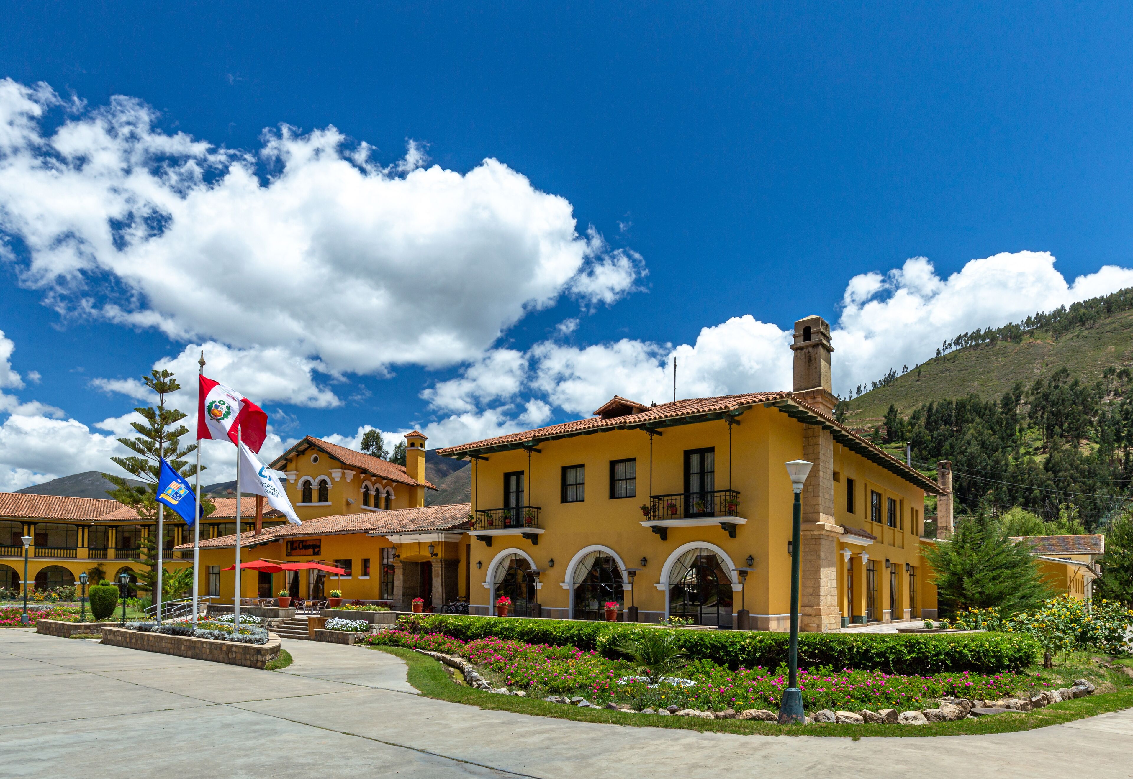 A yellow building with arched windows and a red roof, surrounded by a landscaped garden with flowers and a flagpole.