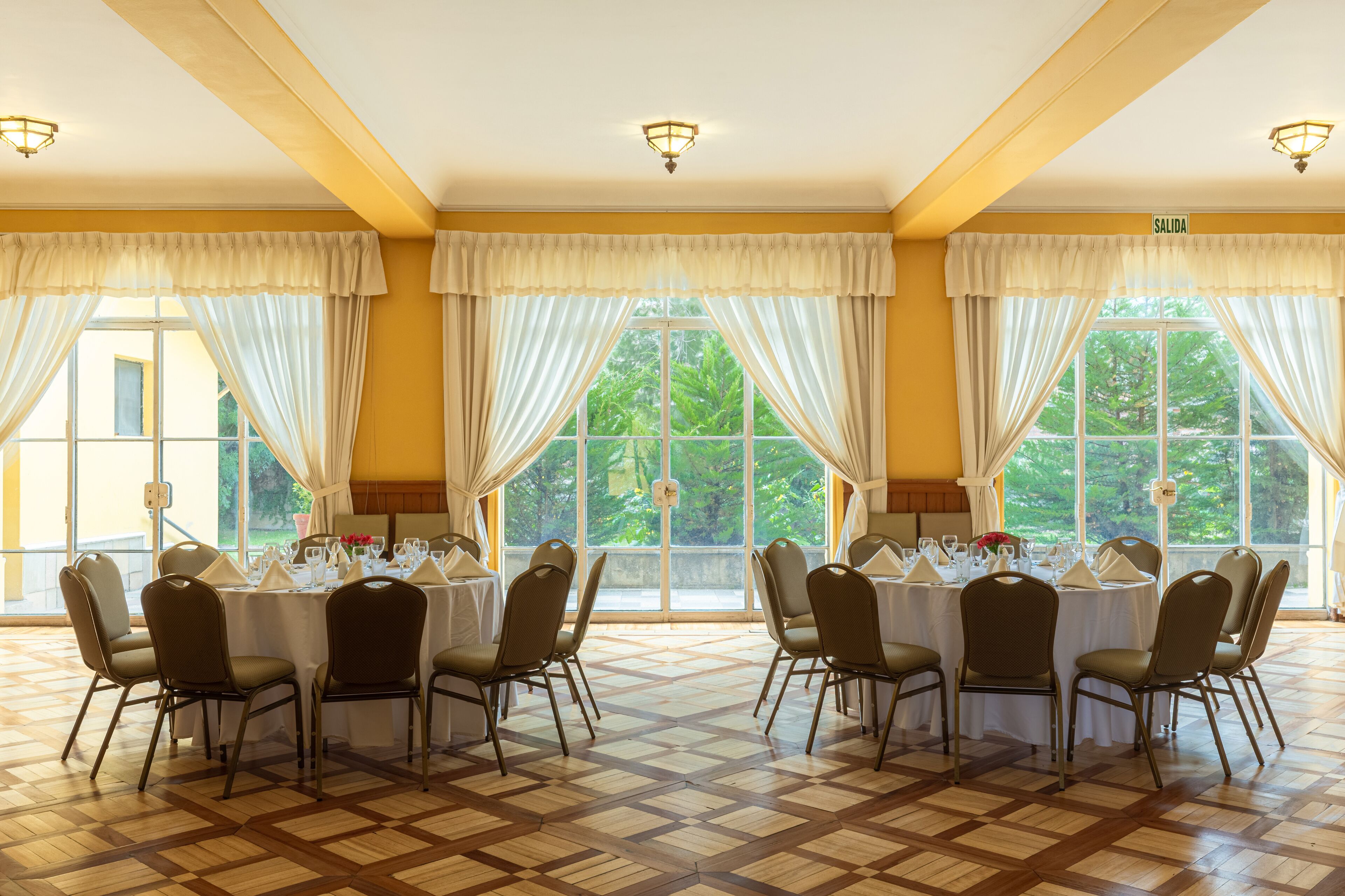 Une salle de conférence dotée d’une grande table préparée pour une réunion, entourée de chaises, de grandes fenêtres avec des rideaux et d’un plancher en parquet.