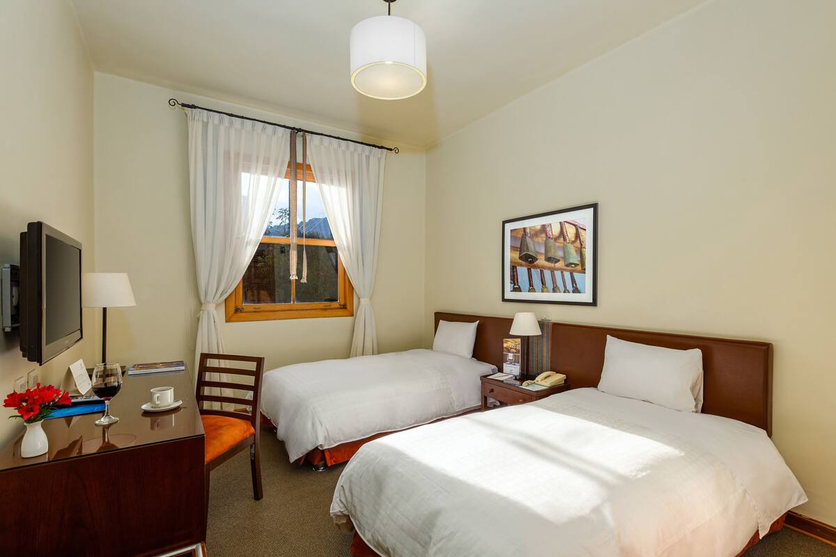 A hotel room with two beds, a desk with a TV, a chair, a window with curtains, and a framed picture on the wall.