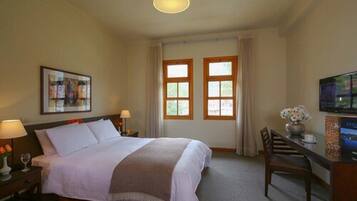 Habitación de hotel con cama, dos ventanas, un escritorio con silla, televisor y un cuadro en la pared.