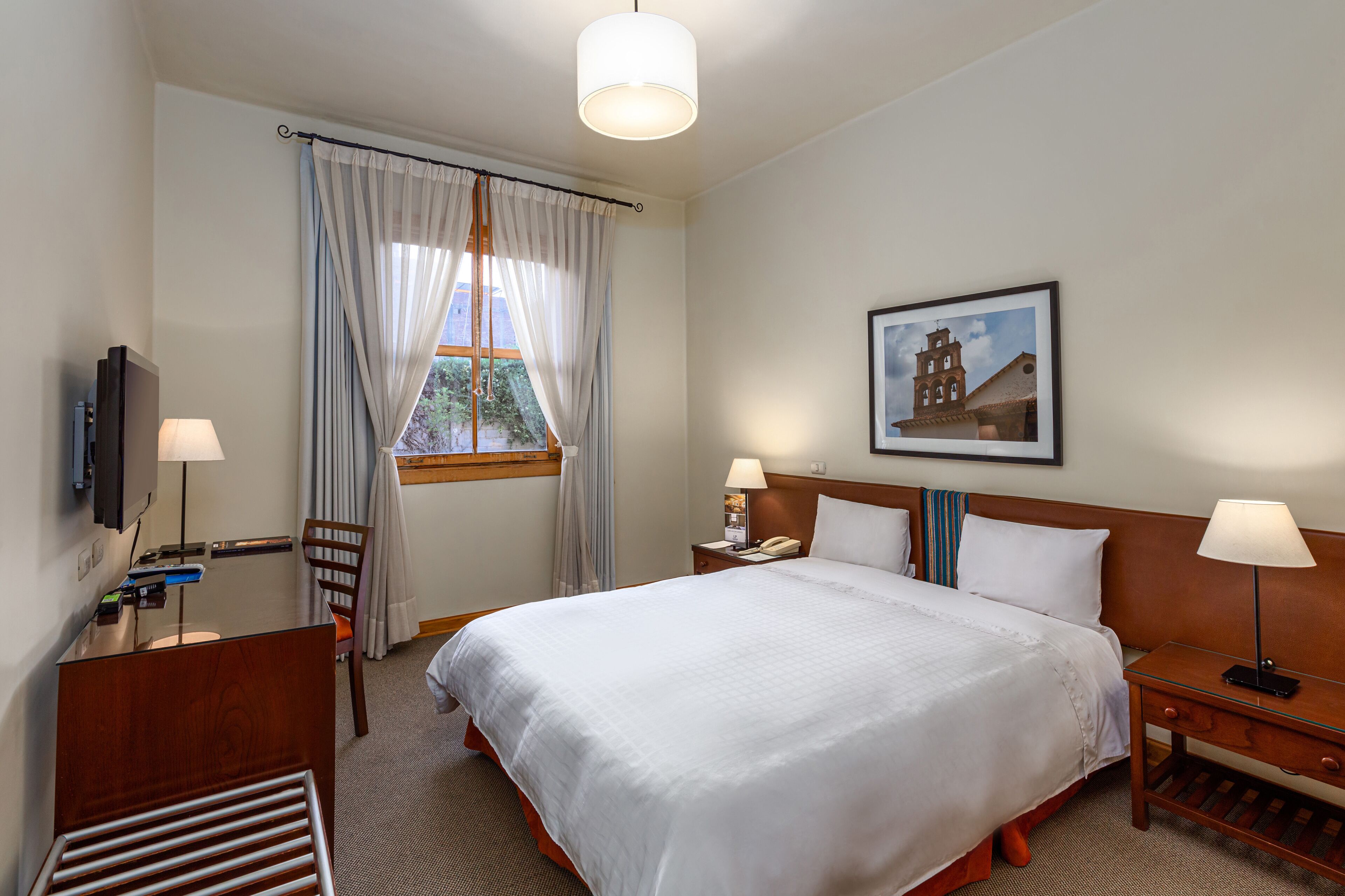 Habitación de hotel con una cama grande, un escritorio, una silla, un televisor, una ventana con cortinas, un cuadro en la pared y una lámpara en la mesita de noche.