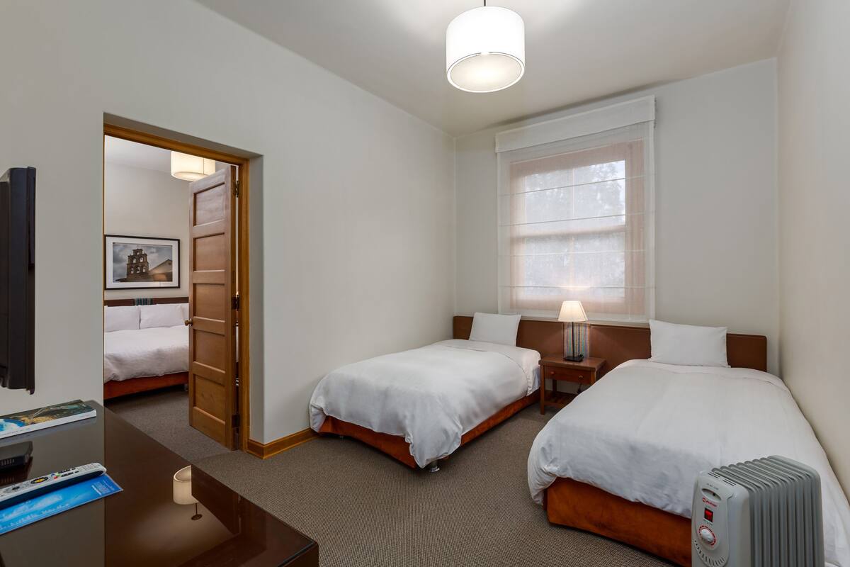 A hotel room with two single beds, a desk with a lamp, a TV, and a heater.