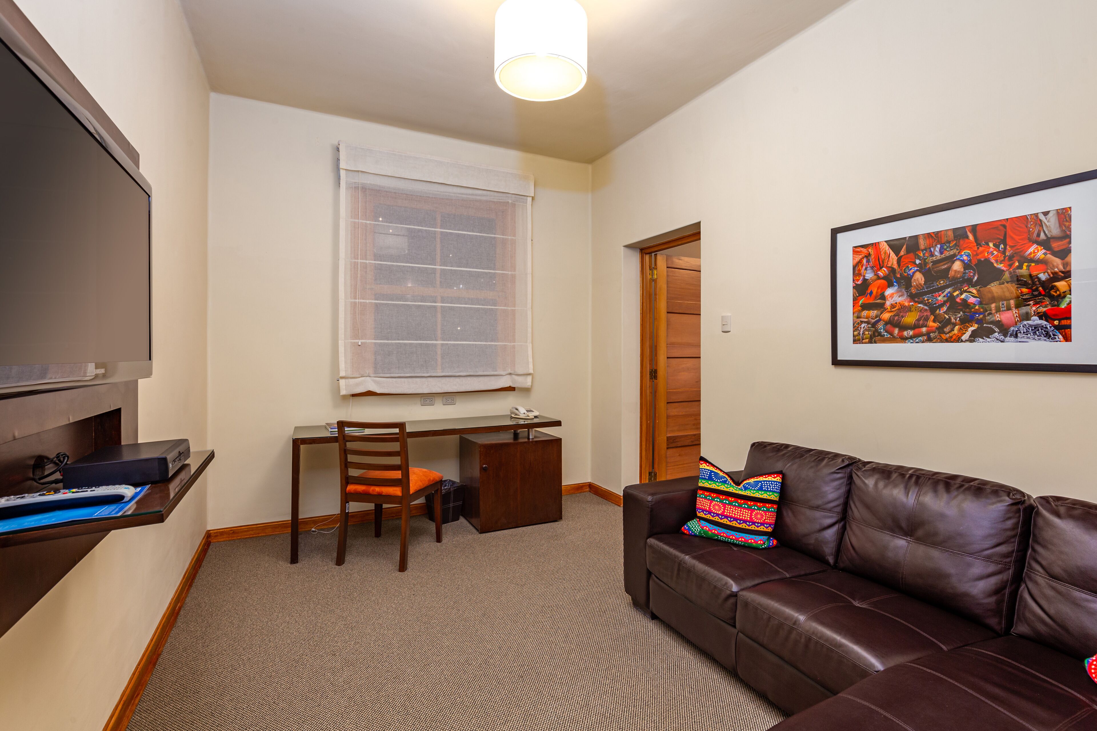 A compact living room with a brown leather sofa, a wooden desk, and a chair. A colorful pillow rests on the sofa.