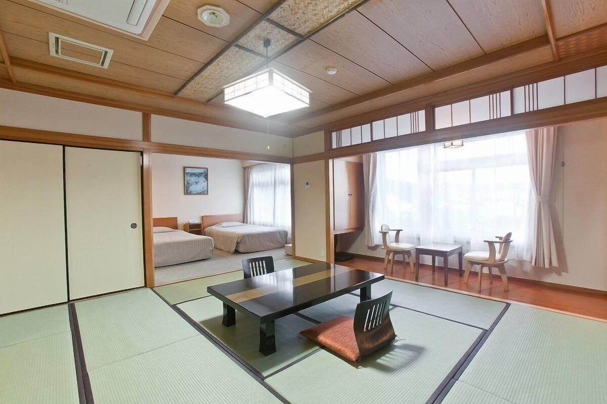 Ein traditioneller japanischer Raum mit Tatami-Böden, einem niedrigen Tisch, Stühlen, einem Bett und einem großen Fenster mit Vorhängen.