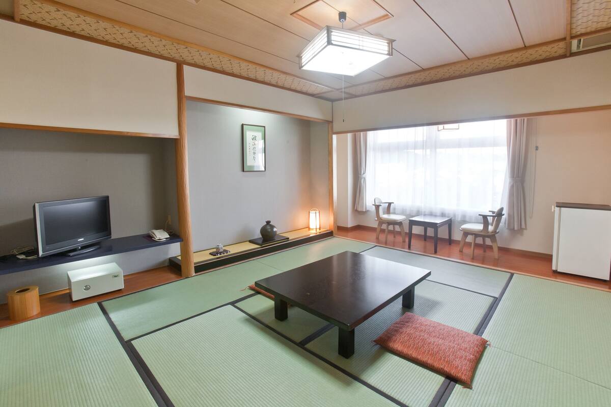 Ein traditioneller japanischer Raum mit Tatami-Böden, einem Fernseher, einem Tisch und einem Fenster mit Vorhängen.
