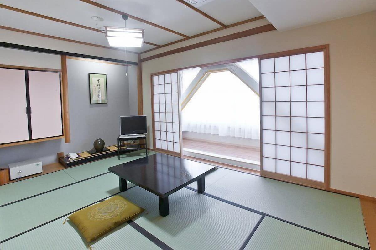 Um quarto japonês tradicional com tatames, uma mesa baixa, portas deslizantes, uma televisão e uma obra de caligrafia emoldurada.