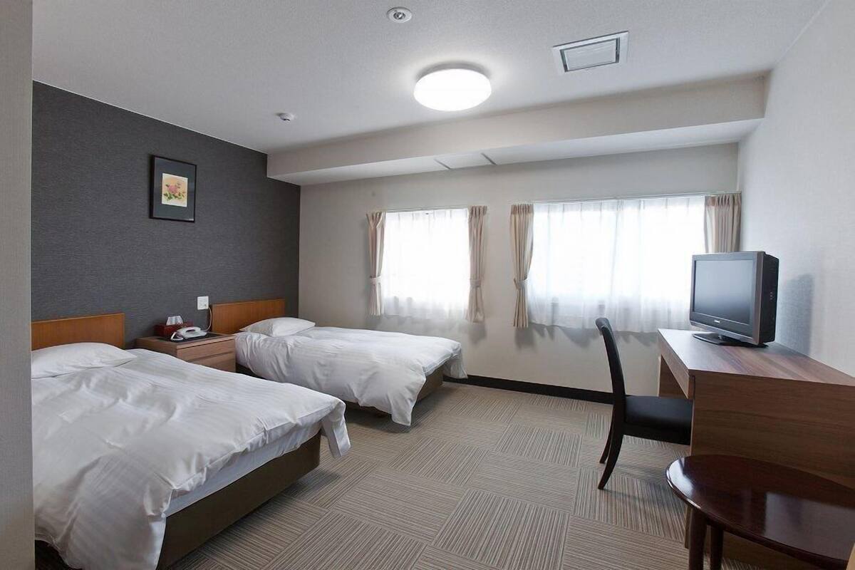 Ein Hotelzimmer mit zwei Betten, einem Schreibtisch, einem Fernseher und einem Fenster mit Vorhängen.