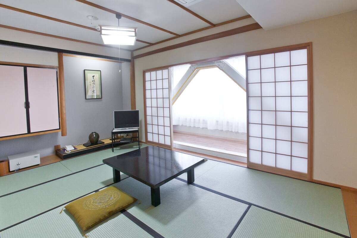 Ein traditioneller japanischer Raum mit Tatami-Böden, einem niedrigen Tisch, einem Fernseher und Schiebetüren.