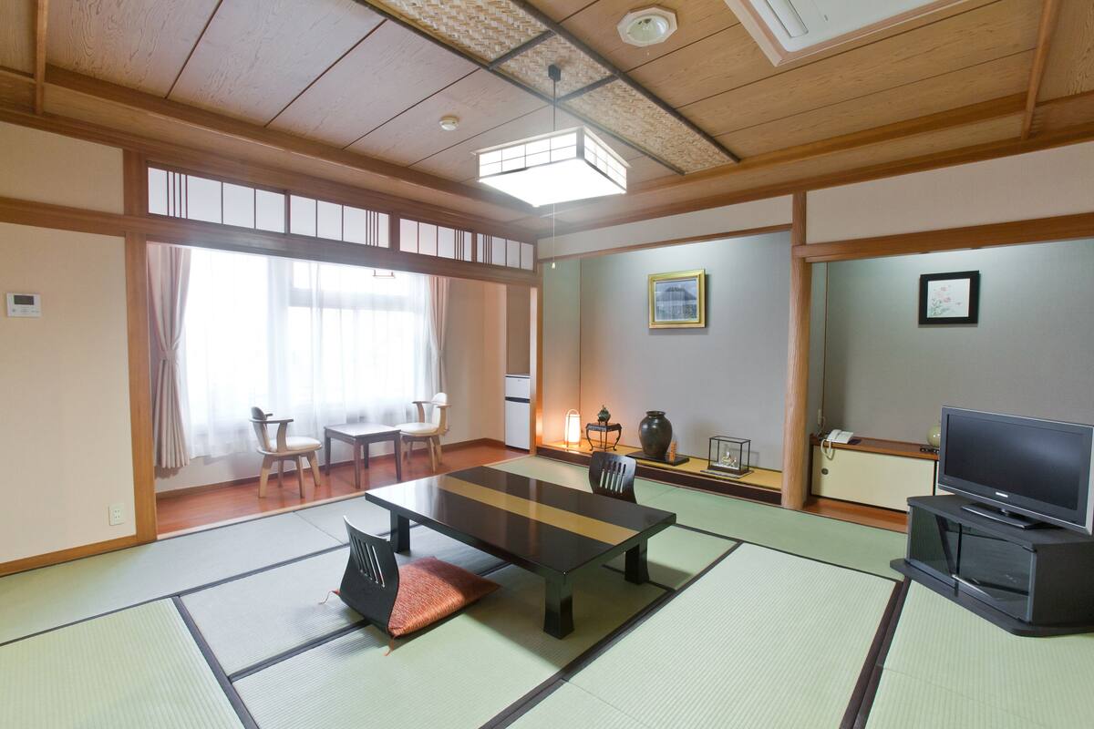 Ein traditioneller japanischer Raum mit Tatami-Böden, einem niedrigen Tisch, Stühlen, einem Fernseher und einer verschiebbaren Tür zu einem weiteren Raum.