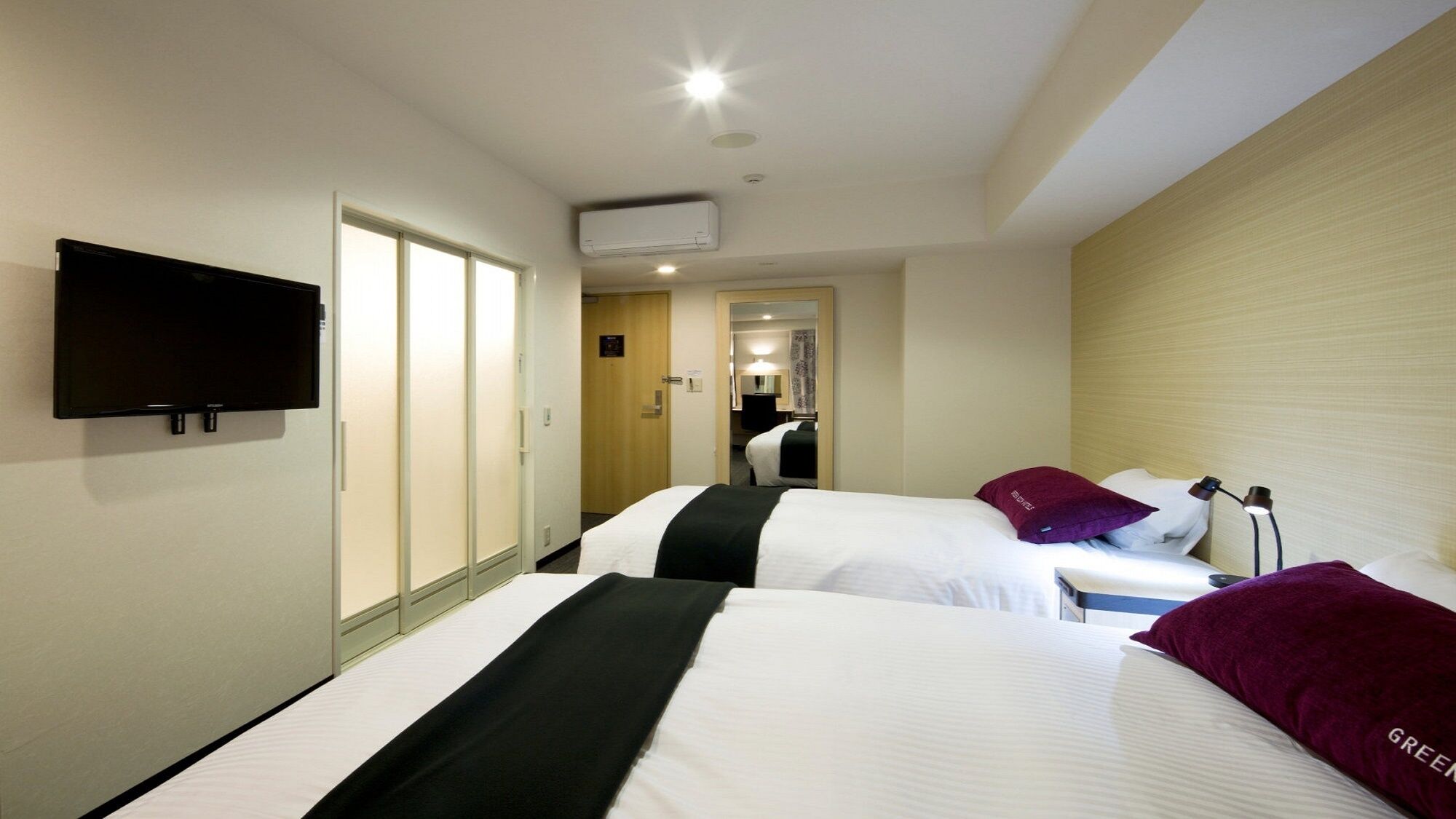 A hotel room with two beds, a TV mounted on the wall, an air conditioning unit, and a door leading to a bathroom.
