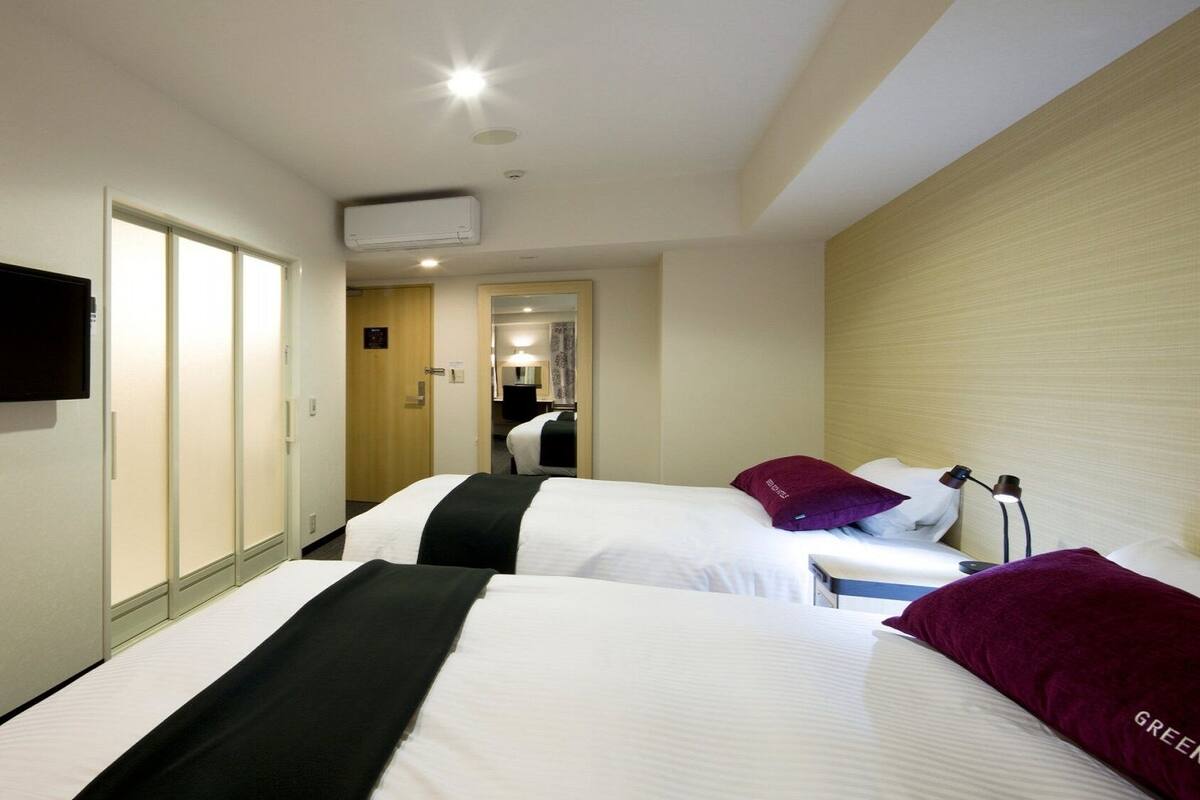 A hotel room with two beds, a TV mounted on the wall, an air conditioning unit, and a door leading to a bathroom.