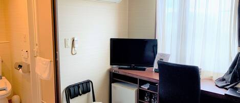 A hotel room with a bed, a desk with a computer, a television, a window with curtains, a bathroom with a toilet, and an air conditioning unit.