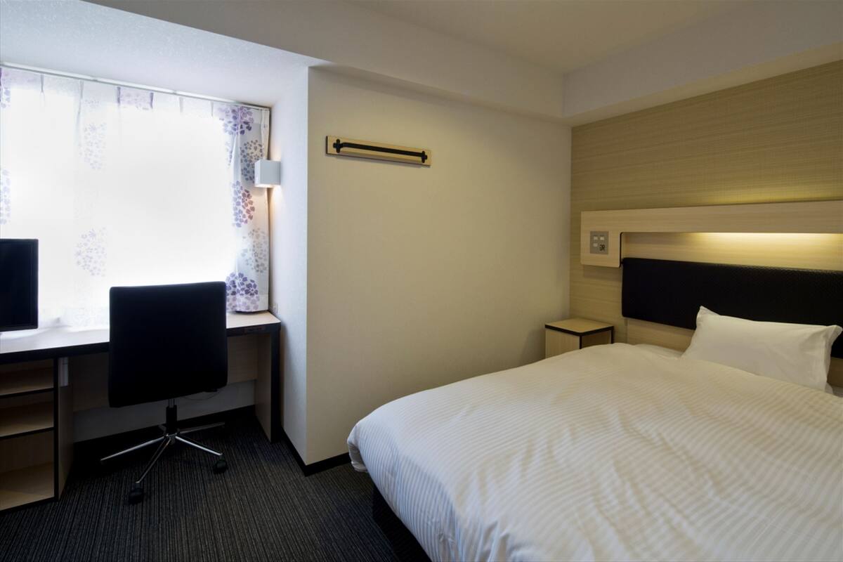 A hotel room with a bed, a desk with a chair, a nightstand, and a window with curtains.