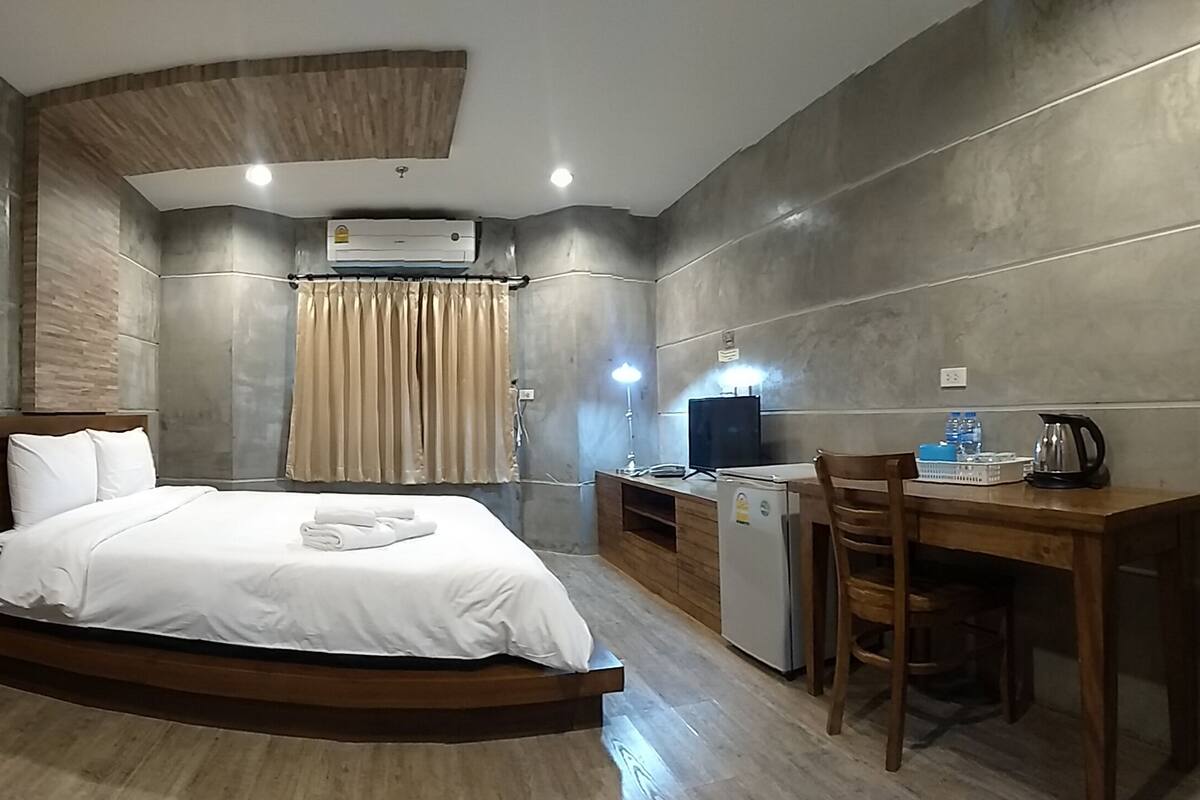 A compact hotel room with a bed, a desk, a chair, a small refrigerator, and an air conditioning unit.