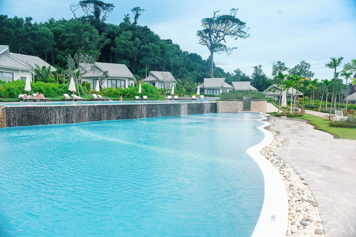 A resort pool with lounge chairs and umbrellas, surrounded by lush greenery and resort buildings.