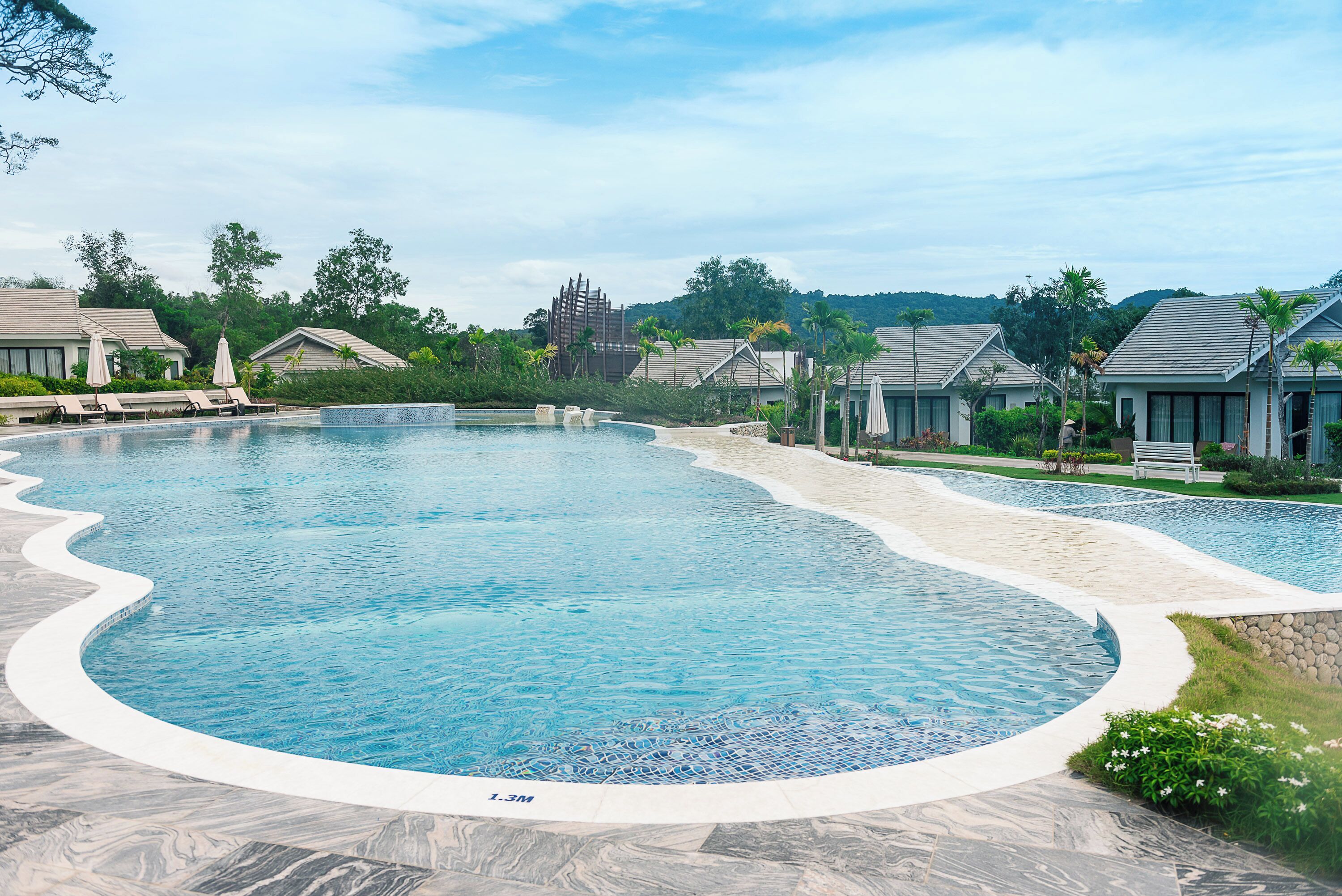 A large, curved swimming pool with a stone border, surrounded by lounge chairs and umbrellas, set in a resort area with villas and greenery.