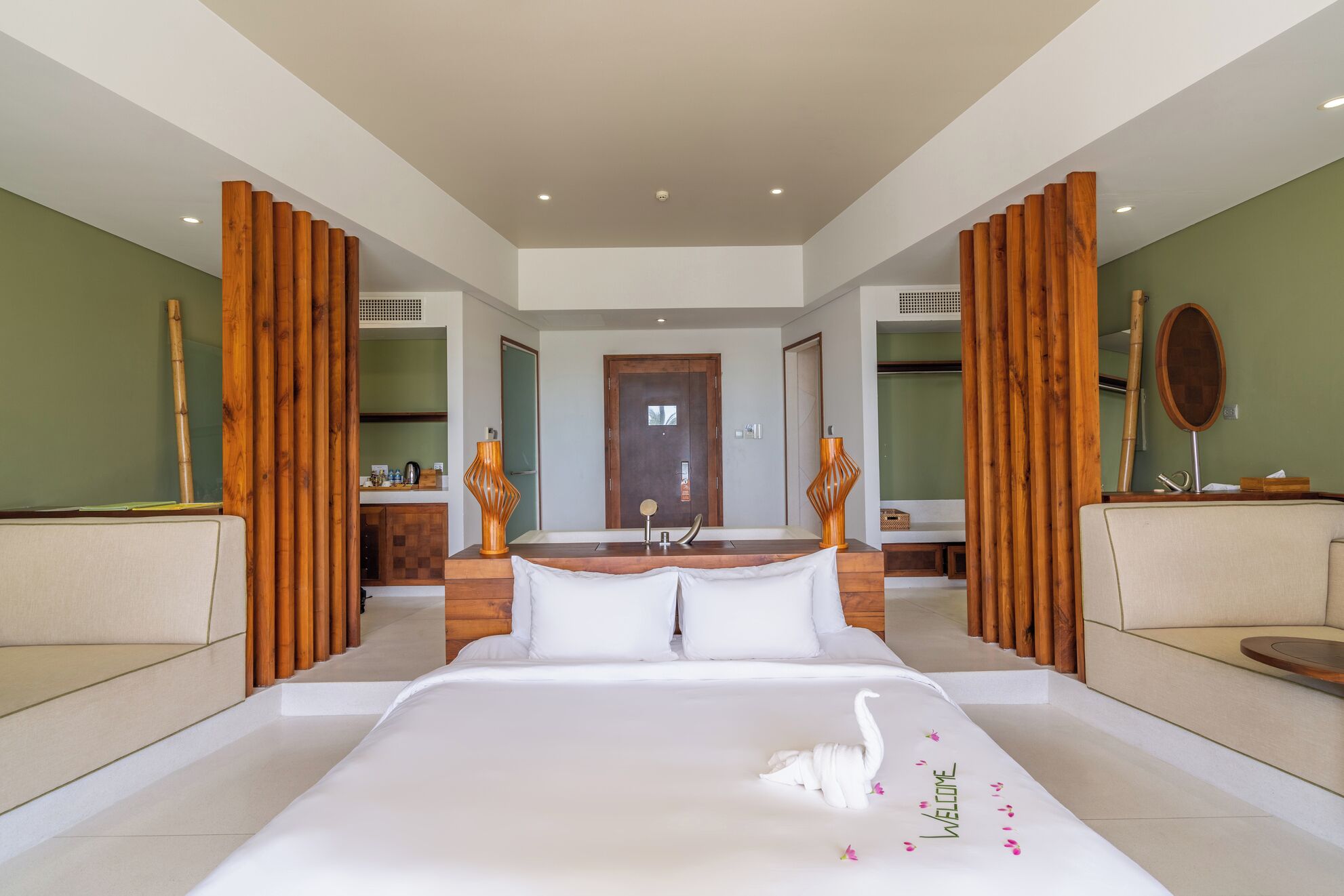 A modern hotel room with a large bed, wooden panels, a bathroom, and a reception area.