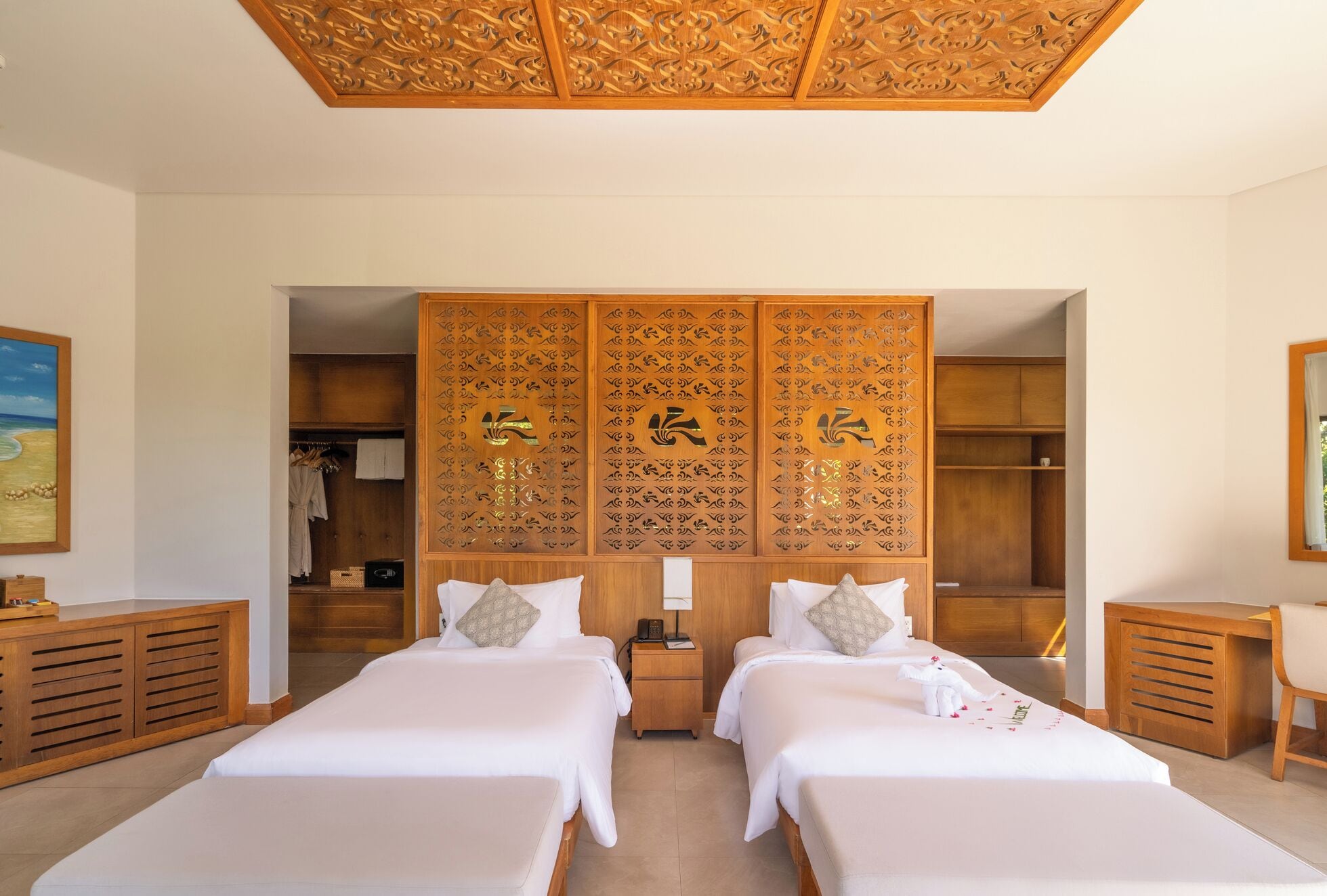 A hotel room with two beds, a wooden wardrobe, a painting on the wall, and a ceiling with intricate carvings.