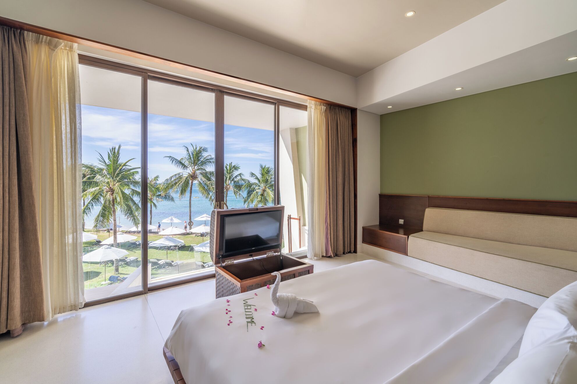 A hotel room with a large bed, a TV on a stand, a sofa, and a view of palm trees and a beach.