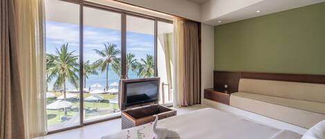 A hotel room with a large bed, a TV on a stand, a sofa, and a view of palm trees and a beach.