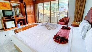 Habitación de hotel con cama, televisión, minibar, armario de madera, una silla roja y balcón con vistas a edificios y vegetación.