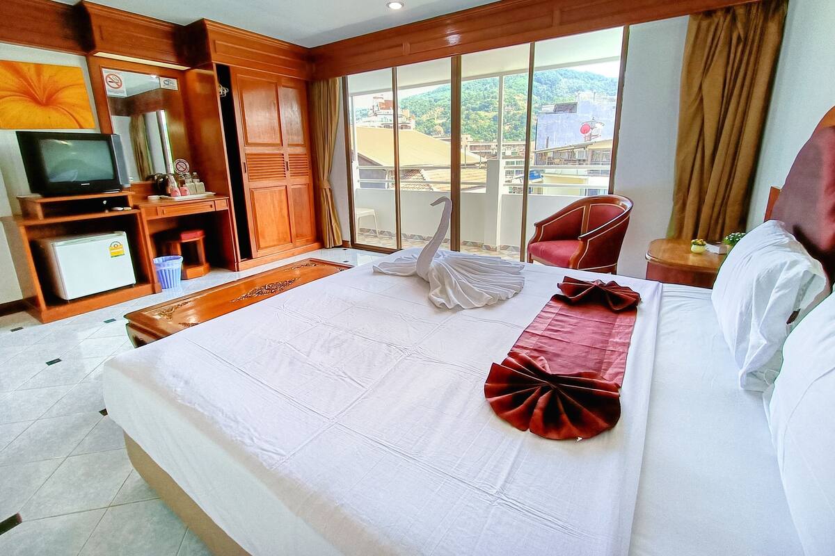 A hotel room with a bed, a television, a mini-fridge, a wooden wardrobe, a red chair, and a balcony with a view of buildings and greenery.