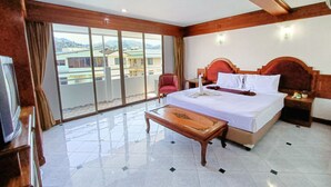 Una habitación de hotel con una cama grande, cabecero de madera, mesita de noche, televisor y balcón con vistas a edificios.
