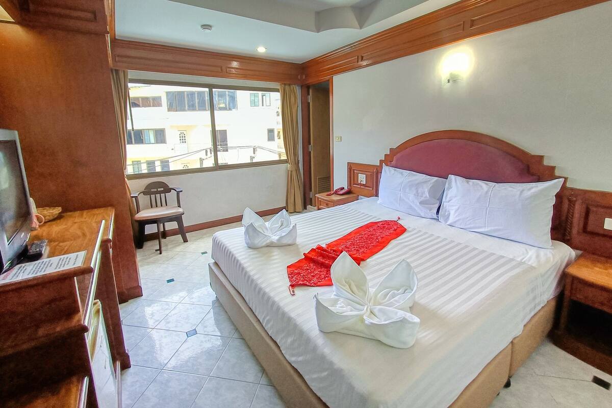 A hotel room with a large bed, a desk with a television, and a window with a view of buildings.