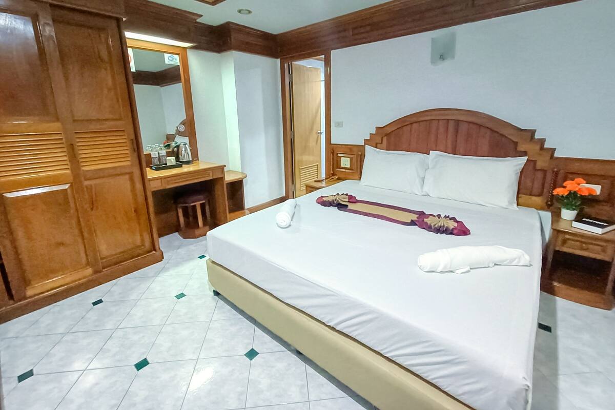 A hotel room with a large bed, wooden headboard, a desk with a chair, a mirror, and a nightstand with a plant.