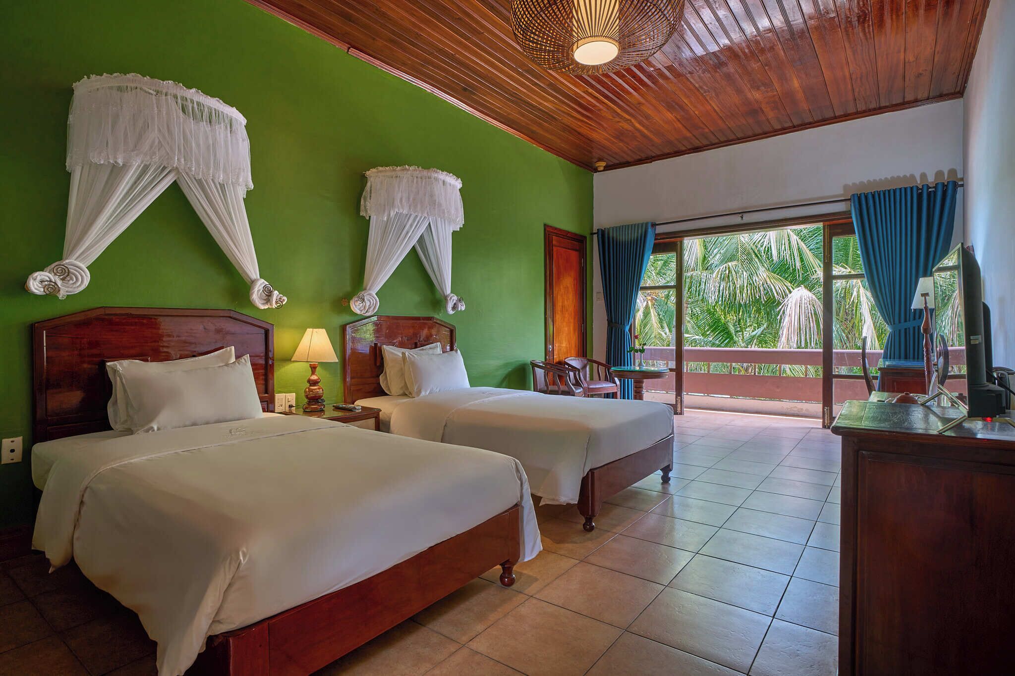 A hotel room with two beds, a wooden ceiling, a green wall, and a view of palm trees.