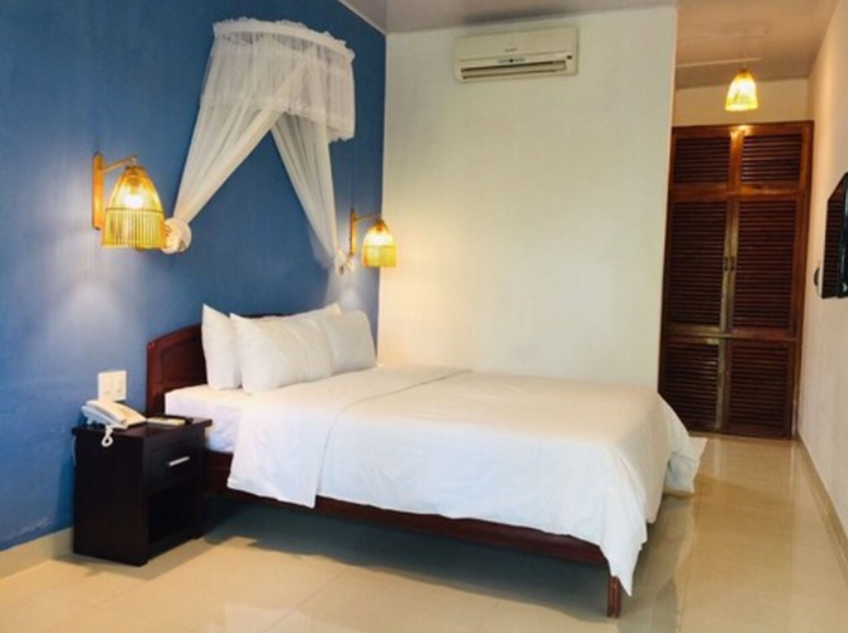 A bedroom with a bed, bedside table, lamp, air conditioning unit, and a door with shutters.